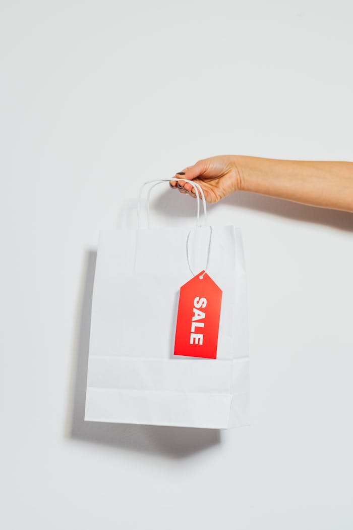 about-us-02 A person's hand holding a white shopping bag with a red sale tag on a white background.